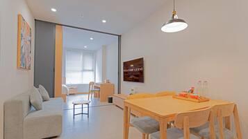 A modern living room with a sofa, dining table, and chairs. There is a television mounted on the wall and a sign that reads "WELCOME GUEST".