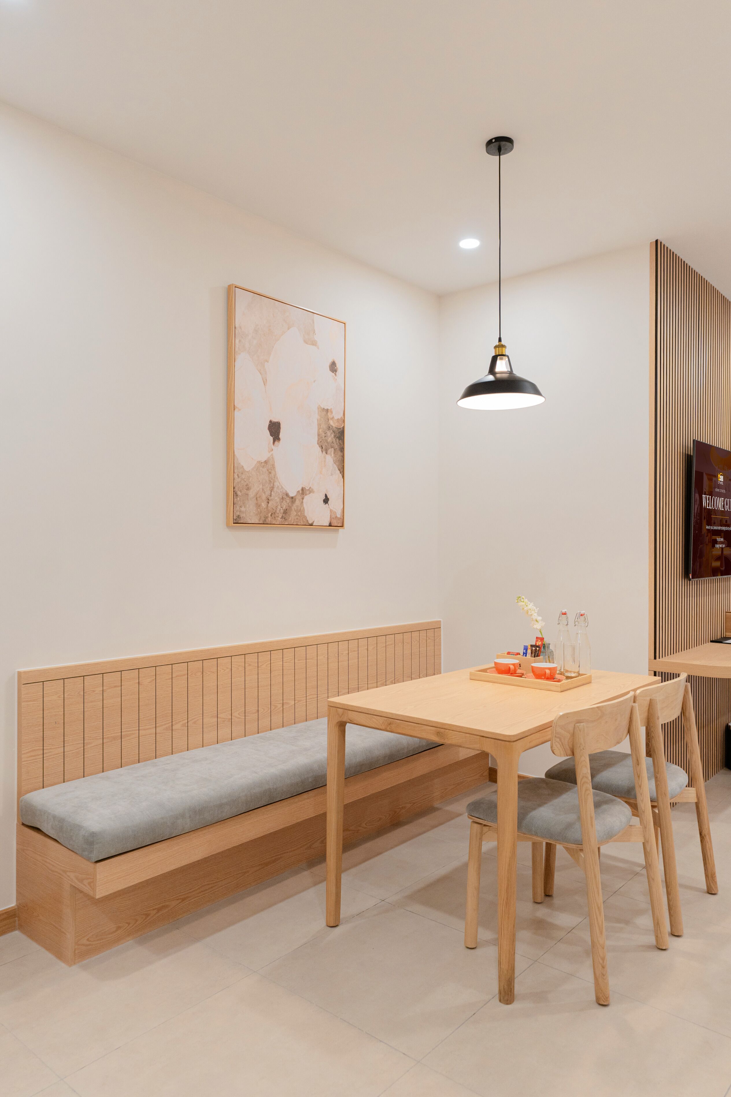 Un comedor moderno con un banco y mesa de madera, una lámpara colgante y un televisor fijado a la pared.