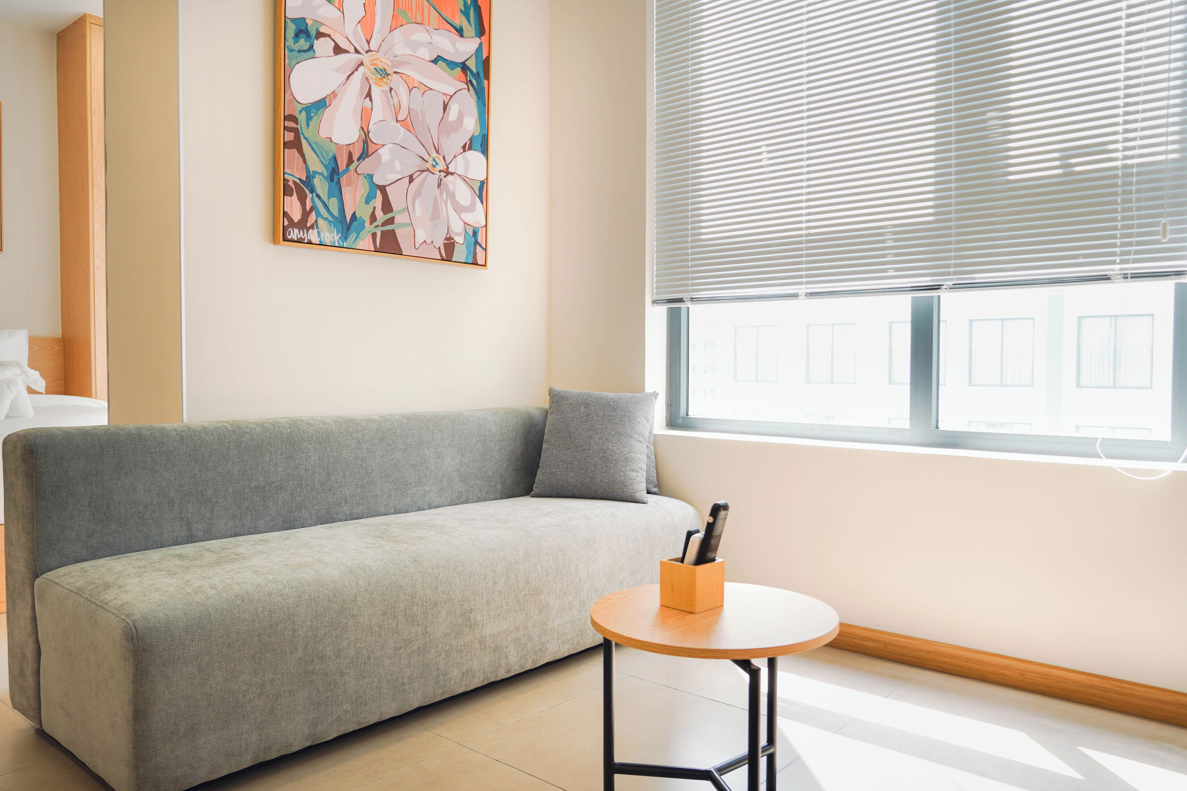 A modern living room with a grey sofa, a small round table with a knife holder, and a large window with blinds.