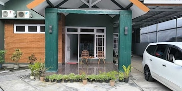 Entrada de um edifício com varanda coberta, duas cadeiras de vime e um carro branco estacionado.