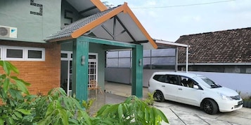 Uma casa com varanda coberta, um carro branco estacionado na frente e um muro de tijolos.