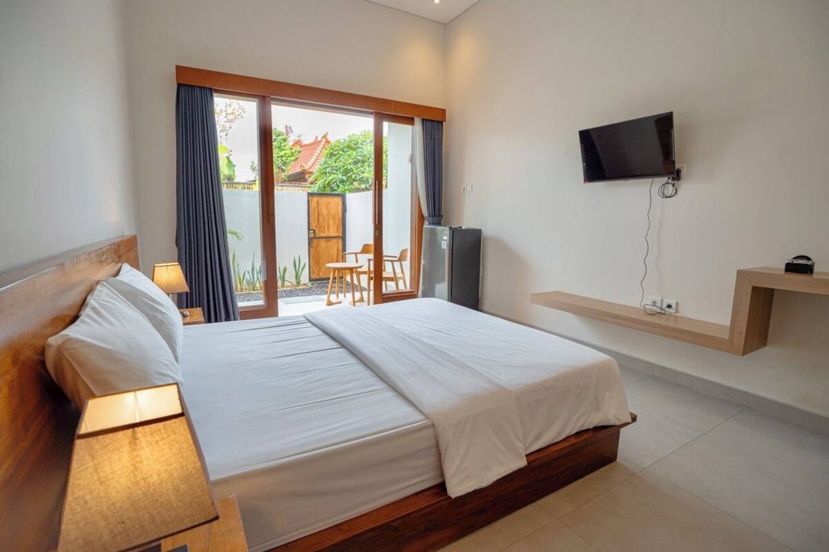 A hotel room with a bed, a desk, a chair, a TV mounted on the wall, and a sliding glass door leading to an outdoor area.