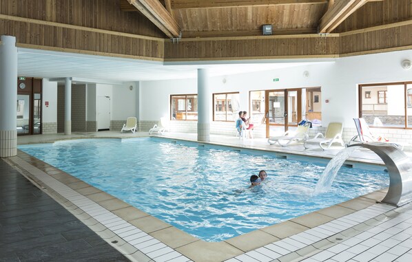 An indoor swimming pool with a wooden ceiling, tiled walls, and lounge chairs around the pool.
