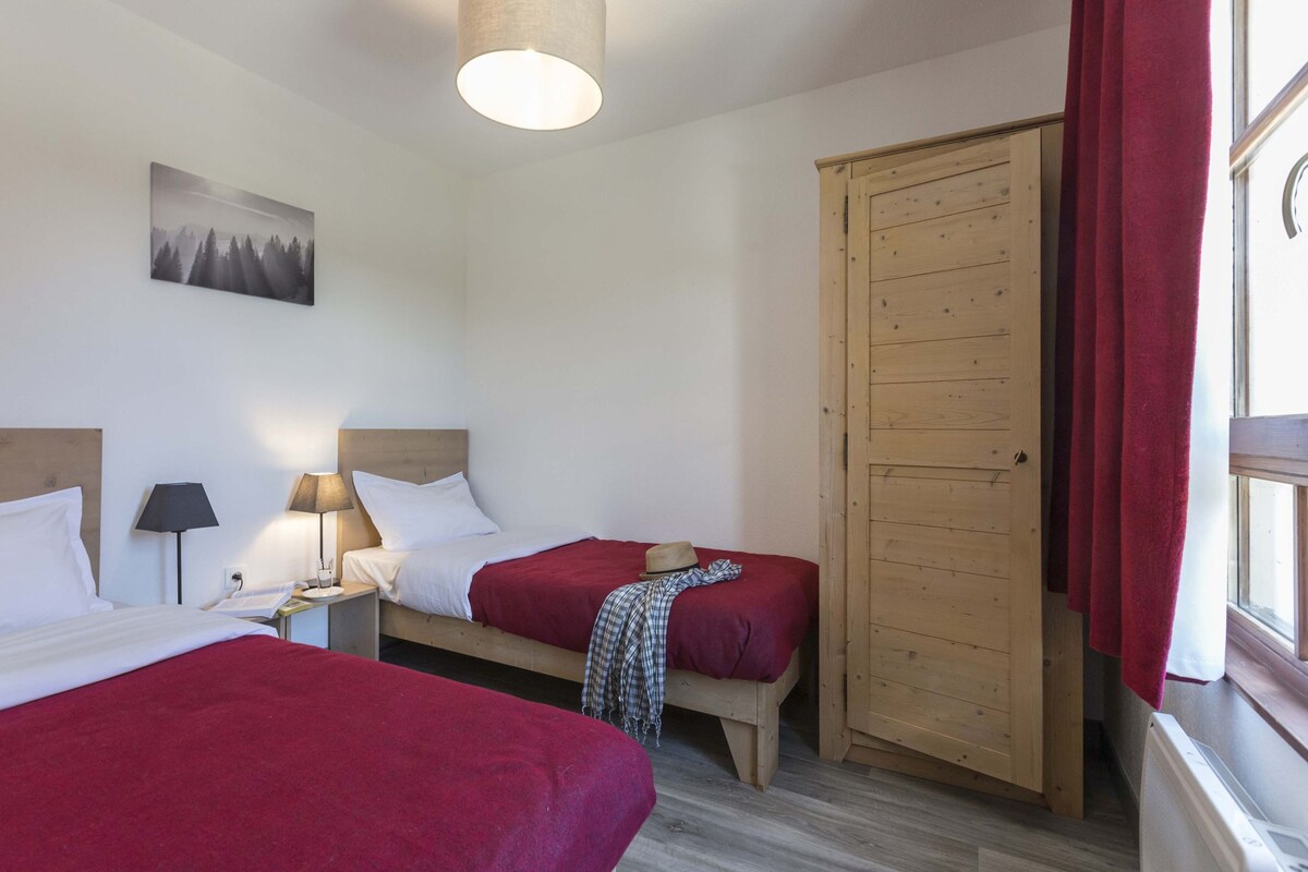 Ein Zimmer mit zwei Betten, einem Holzschrank, einem Fenster mit roten Vorhängen, einer Lampe und einem Wandbild.