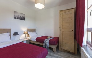 A room with two beds, a wooden wardrobe, a window with red curtains, a lamp, and a wall-mounted picture.