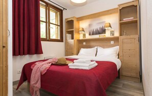 A bedroom with a bed, a wooden wardrobe, a window with curtains, and a wall-mounted lamp.