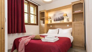 A bedroom with a bed, a wooden wardrobe, a window with curtains, and a wall-mounted lamp. - Résidence Odalys L'orée des Pistes (Saint-Sorlin-d'Arves)