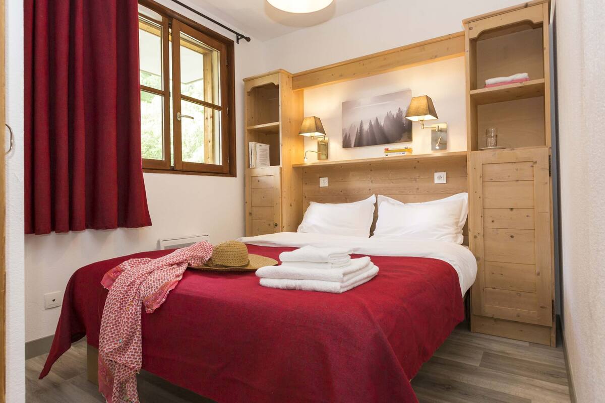 Un dormitorio con cama, armario de madera, ventana con cortinas y lámpara fijada a la pared.