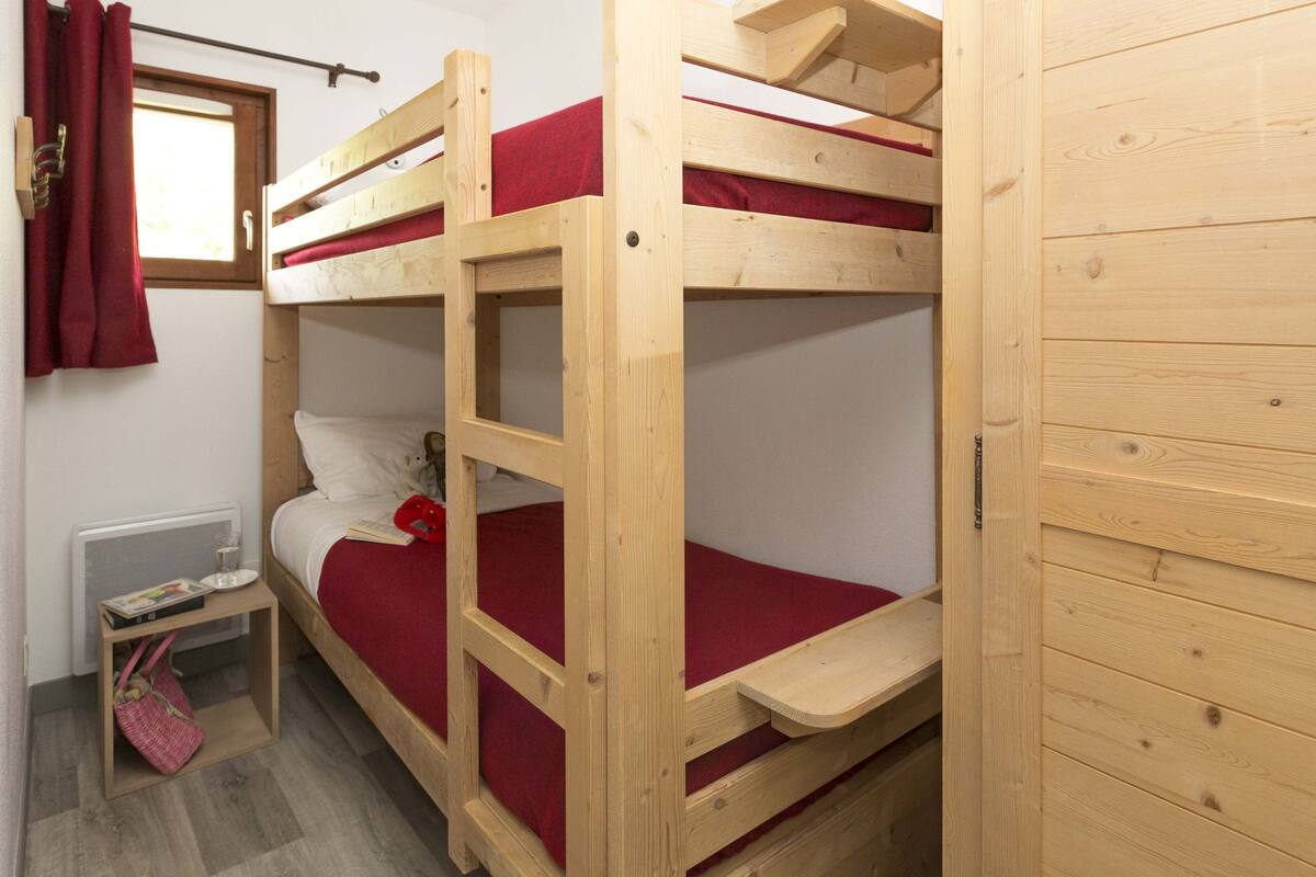 Habitación con literas, una ventana, una mesa pequeña con un libro y una cortina roja.