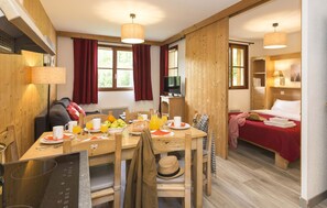 A room with a wooden dining table set for breakfast, a bed with a red blanket, and a television on a wooden stand.