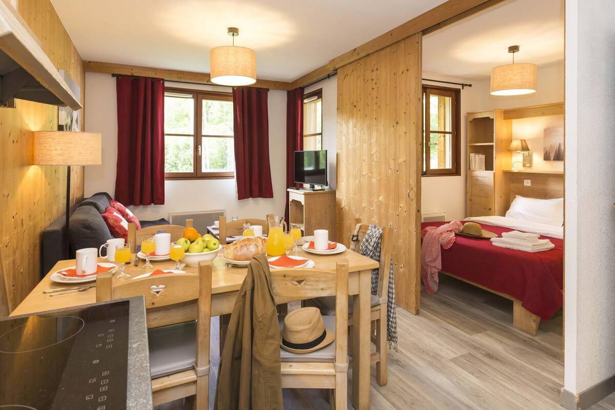 Una habitación con una mesa de madera dispuesta para el desayuno, una cama con una colcha roja y un televisor sobre un mueble de madera.