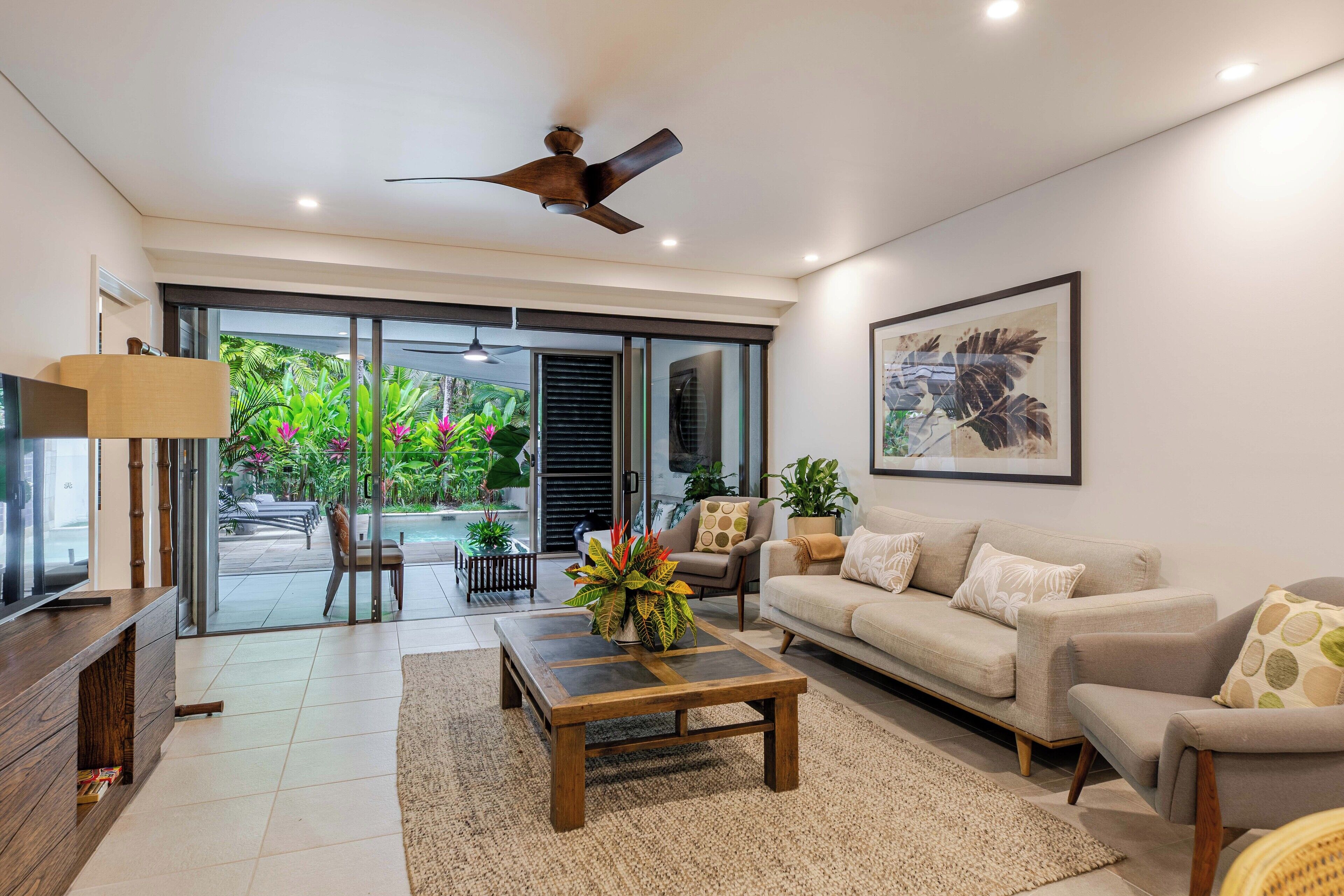 A modern living room with a sofa, coffee table, and TV stand. There is a ceiling fan, a large window with a view of greenery, and a framed picture on the wall.