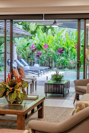 A modern indoor space with a glass wall offering a view of an outdoor garden area with tropical plants and a pool.