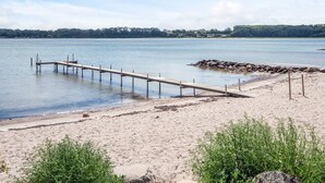 Una playa arenosa con un muelle de madera que se adentra en aguas tranquilas, rodeada de vegetación y un cielo despejado.
