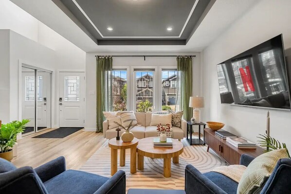 A modern living room with a sofa, two coffee tables, a TV, and a view of houses through large windows.