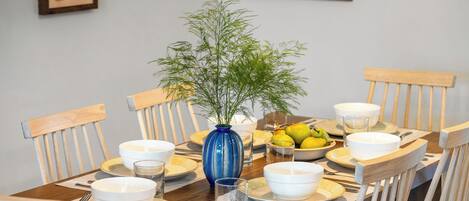 Ein Esstisch, gedeckt mit weißen Schüsseln, Tellern und Besteck, eine blaue Vase mit Grünpflanzen und eine Obstschale.