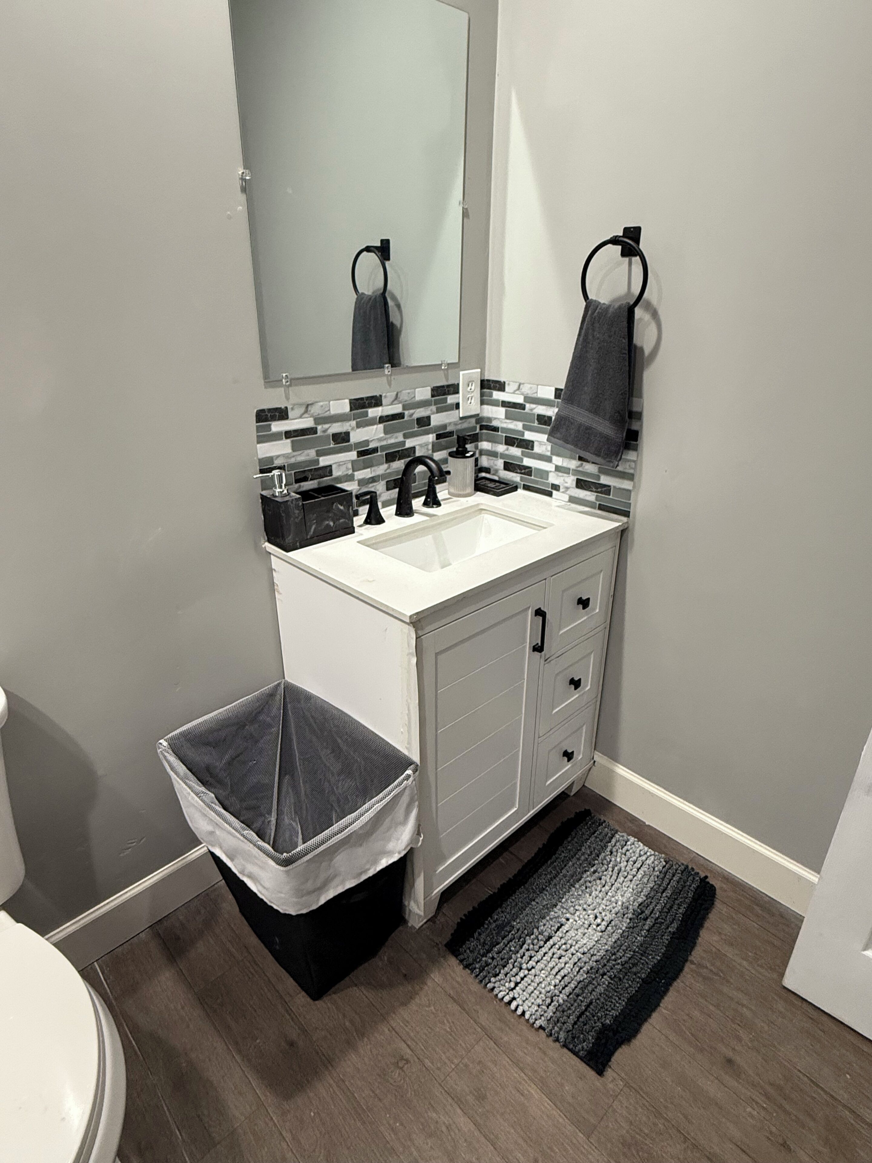 Une salle de bain avec un lavabo blanc, un robinet noir et un mur recouvert de carreaux. On y trouve une poubelle noire et un tapis gris sur le sol.