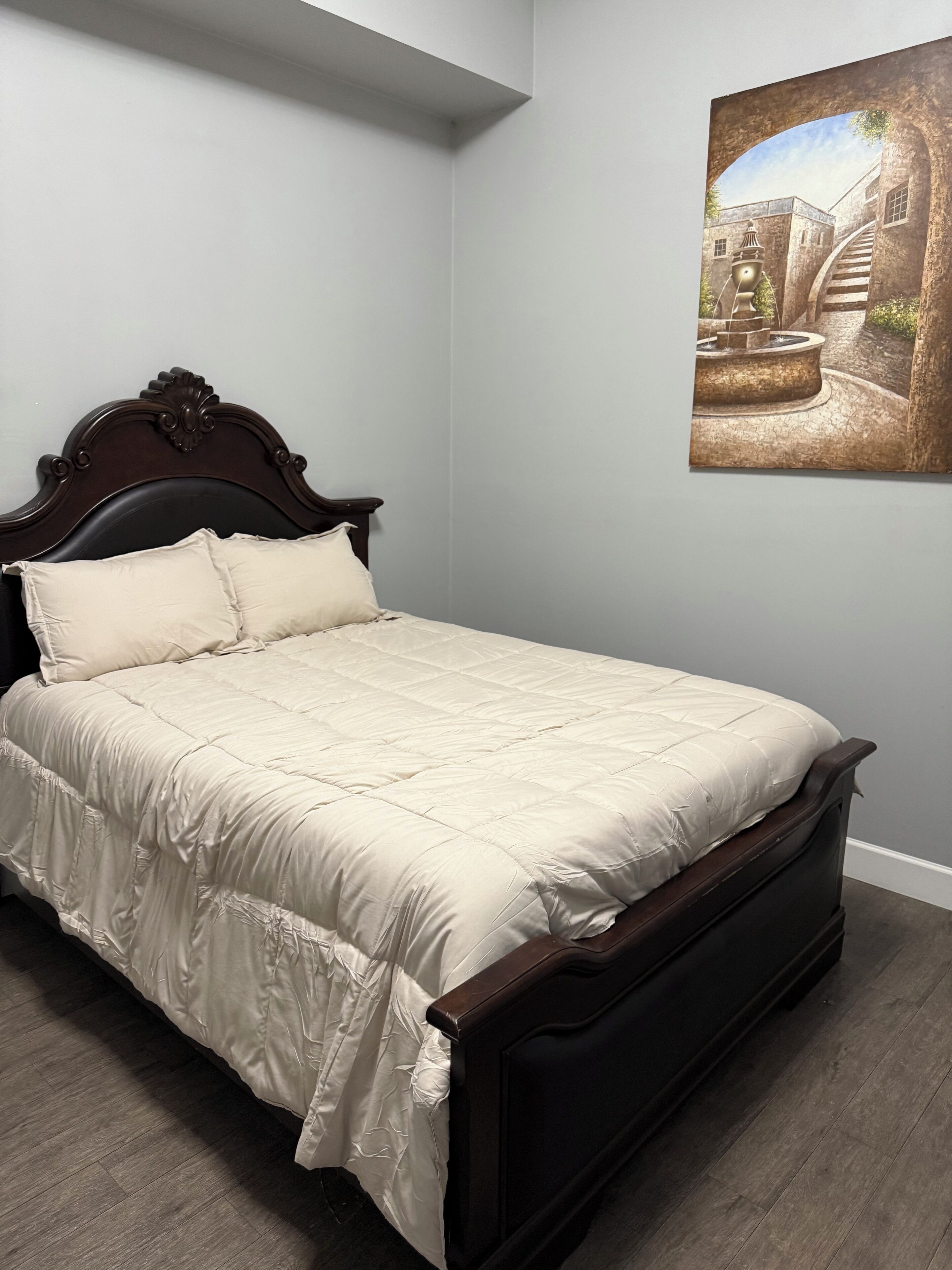 Una cama con cabecera de madera oscura y colcha color crema en una habitación con paredes grises claras y suelo de madera.