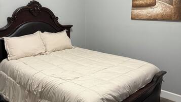 Una cama con cabecera de madera oscura y colcha color crema en una habitación con paredes grises claras y suelo de madera.