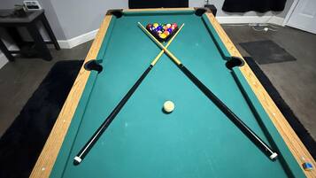 Une table de billard avec une boule blanche et un jeu de billes, une petite table avec un ordinateur portable et une tasse, et une affiche accrochée au mur.