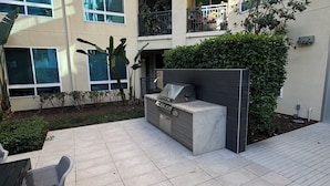 Une cour avec un espace barbecue, des sièges extérieurs et une grande haie.