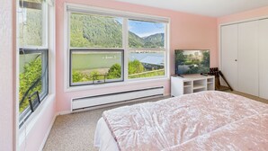 A bedroom with a large window offering a mountain view, a bed with a pink bedspread, a TV on a white stand, and a wardrobe.
