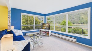 A room with a blue wall, a large window, a sofa, a coffee table, and a TV.