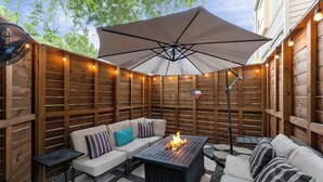 An outdoor patio with a wooden fence, a black table with a fire feature, and a beige sofa with striped pillows. - EaDo Escape • Sleeps 10 - Private Yard & Driveway + Rooftop City Views (Houston)