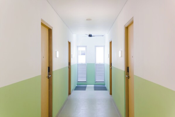 Un pasillo estrecho con dos puertas a cada lado, un ventilador de techo y una ventana con persianas al final.