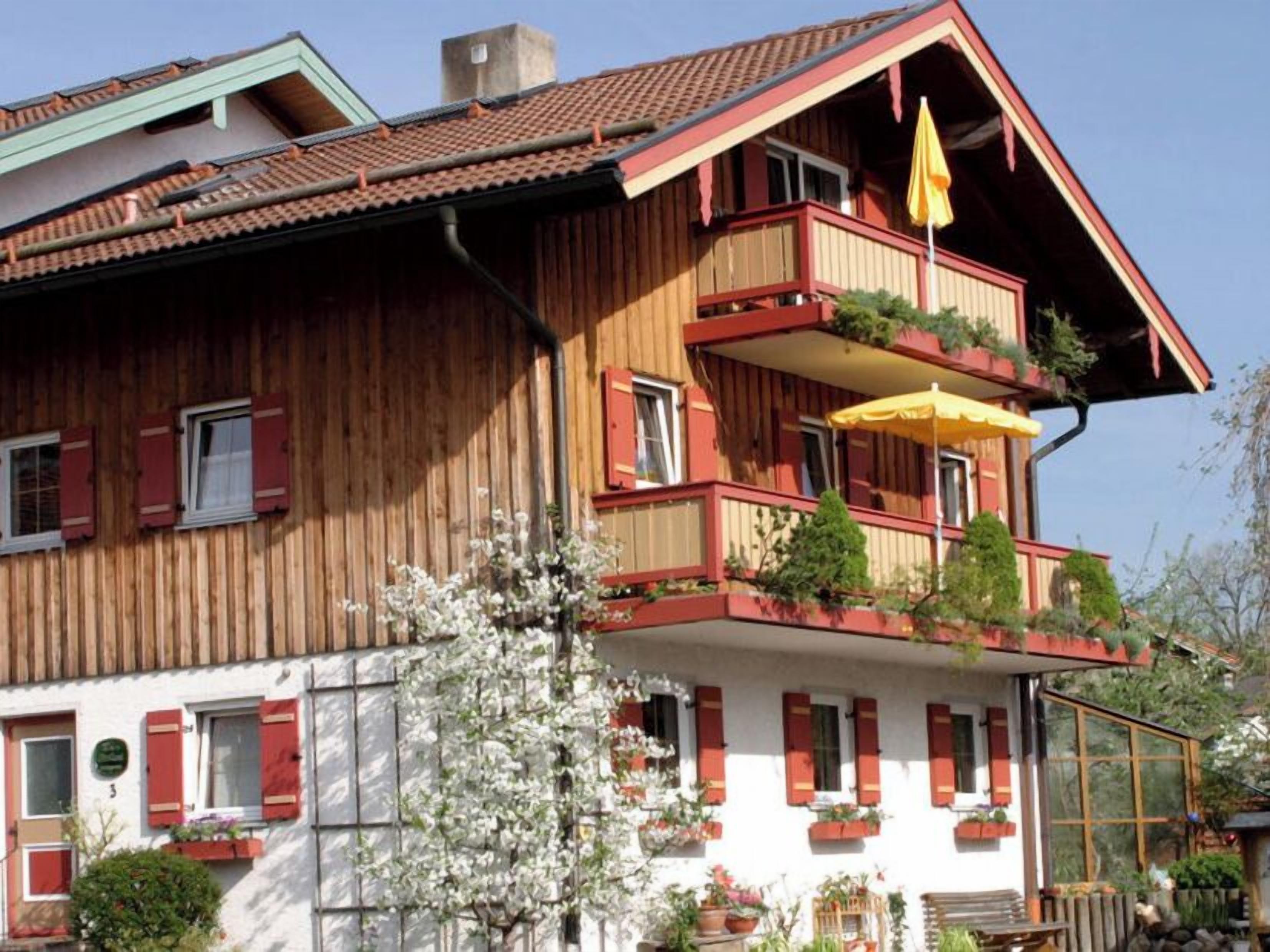 Ein zweistöckiges Holzhaus mit roten Fensterläden, Balkonen mit Pflanzen und einem gelben Sonnenschirm.