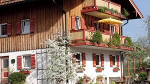 A two-story wooden building with red shutters, balconies with plants, and a yellow umbrella. - Vacation apartment, 58 sqm 1st floor, 2 separate bedrooms (Bad Endorf)