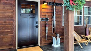 Una cabaña de madera con puerta frontal oscura, esquíes y tableros de snowboard colgados en la pared, un adorno navideño y un banco de madera en el exterior.