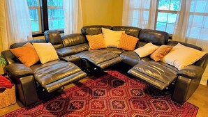 Un sofá modular con varios cojines, una alfombra roja estampada y amplias ventanas con cortinas.