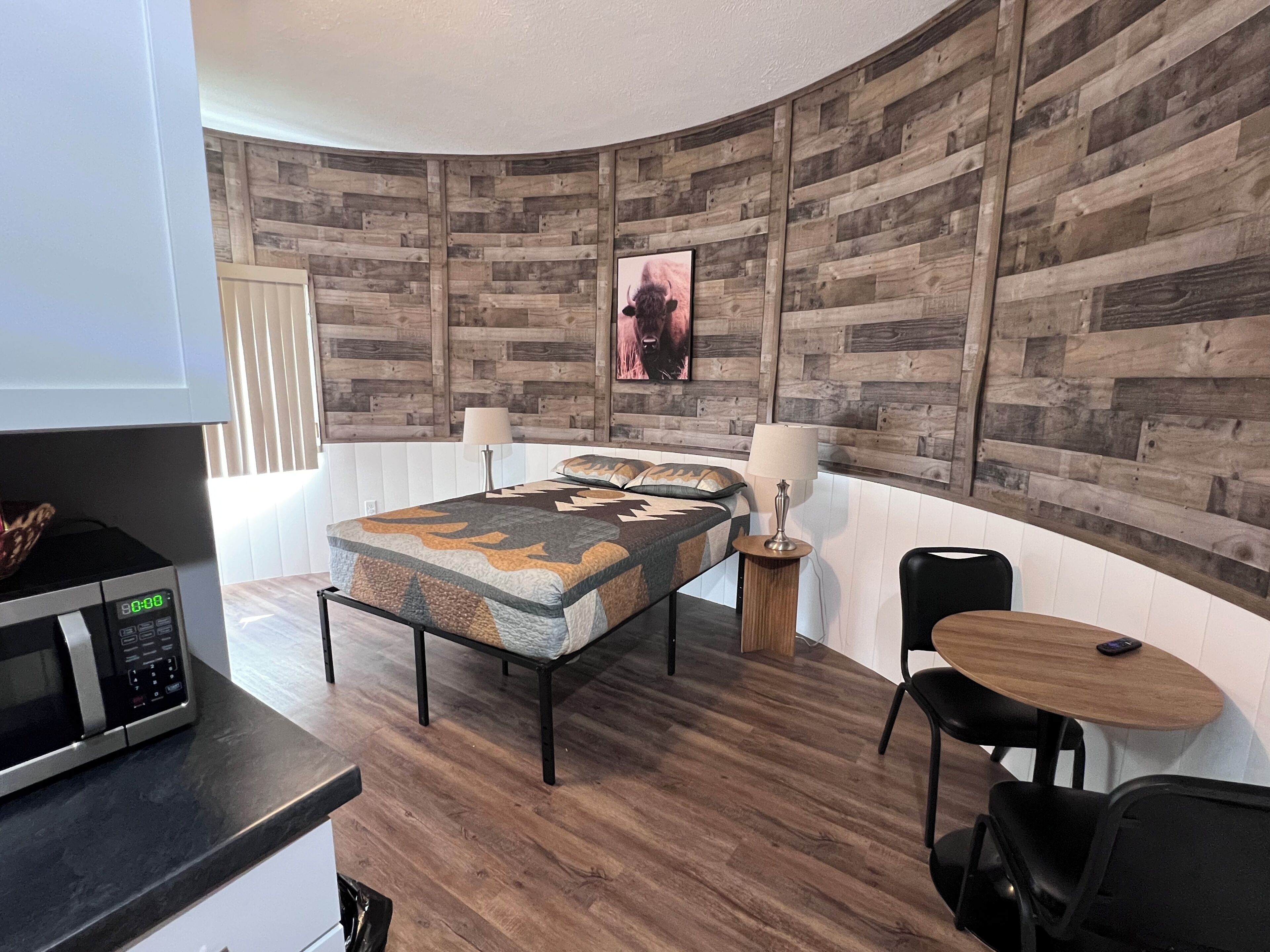 Une chambre moderne avec un panneau mural en bois, un lit recouvert d’une couvre-lit à motifs, une petite table ronde avec une chaise et un four à micro-ondes posé sur un comptoir.