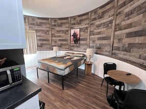 A modern room with a wooden wall panel, a bed with a patterned bedspread, a small round table with a chair, and a microwave on a counter.