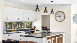A modern kitchen with white cabinets, a large island, pendant lighting, a clock, and a window with a view of trees. - Forest Edge Daylesford (Eganstown)