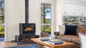 A modern living room with a wood-burning stove, a leather sofa, a coffee table with books, and large windows offering a view of the outdoors. - Forest Edge Daylesford (Eganstown)