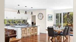 A modern kitchen with white cabinets, a central island with stools, a dining area with a table and chairs, and a large window with a view of trees. - Forest Edge Daylesford (Eganstown)
