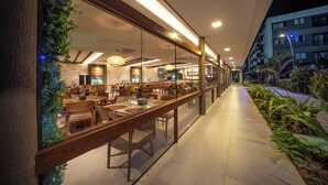 An indoor restaurant with wooden tables and chairs, a glass railing, and a view of the street outside. - Cozy home with Wi-Fi, air conditioning and great location in Muro Alto/PE (Ipojuca)