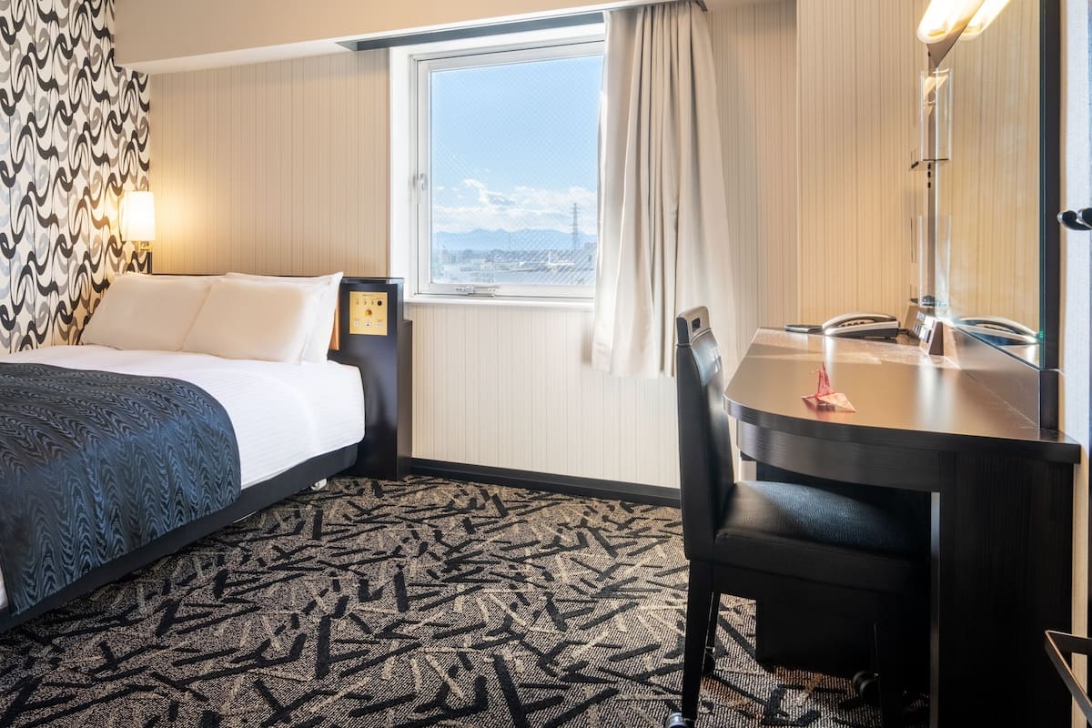 A hotel room with a bed, a desk, a chair, a window with a view, and a patterned carpet.