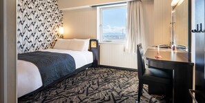 A hotel room with a bed, a desk, a chair, a window with a view, and a patterned carpet.