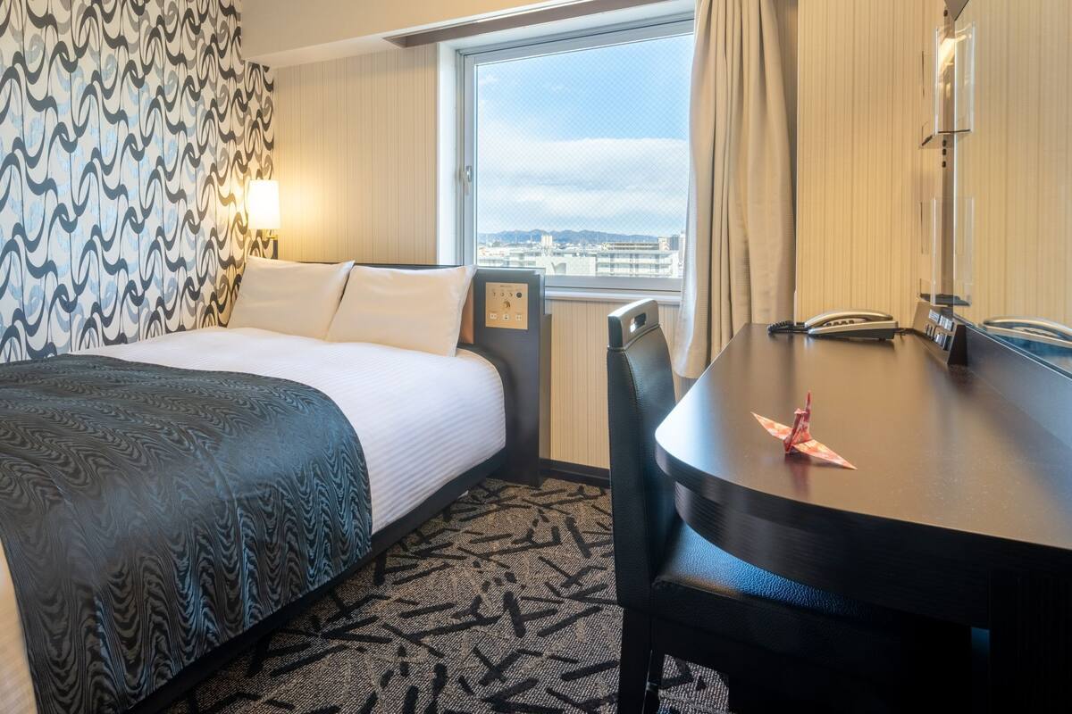 A hotel room with a bed, a desk, a chair, a lamp, a telephone, and a window with a view.