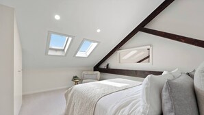 Una camera da letto con soffitto spiovente, lucernari, un letto con biancheria bianca, un tavolino rotondo con una pianta e una sedia con cuscini a righe.