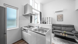 A compact kitchen with white cabinetry, a stainless steel sink, and a built-in stove. There is a grey sofa and a small table with a plant in the living area. - FURANO RESORT WAKABA D (Furano)