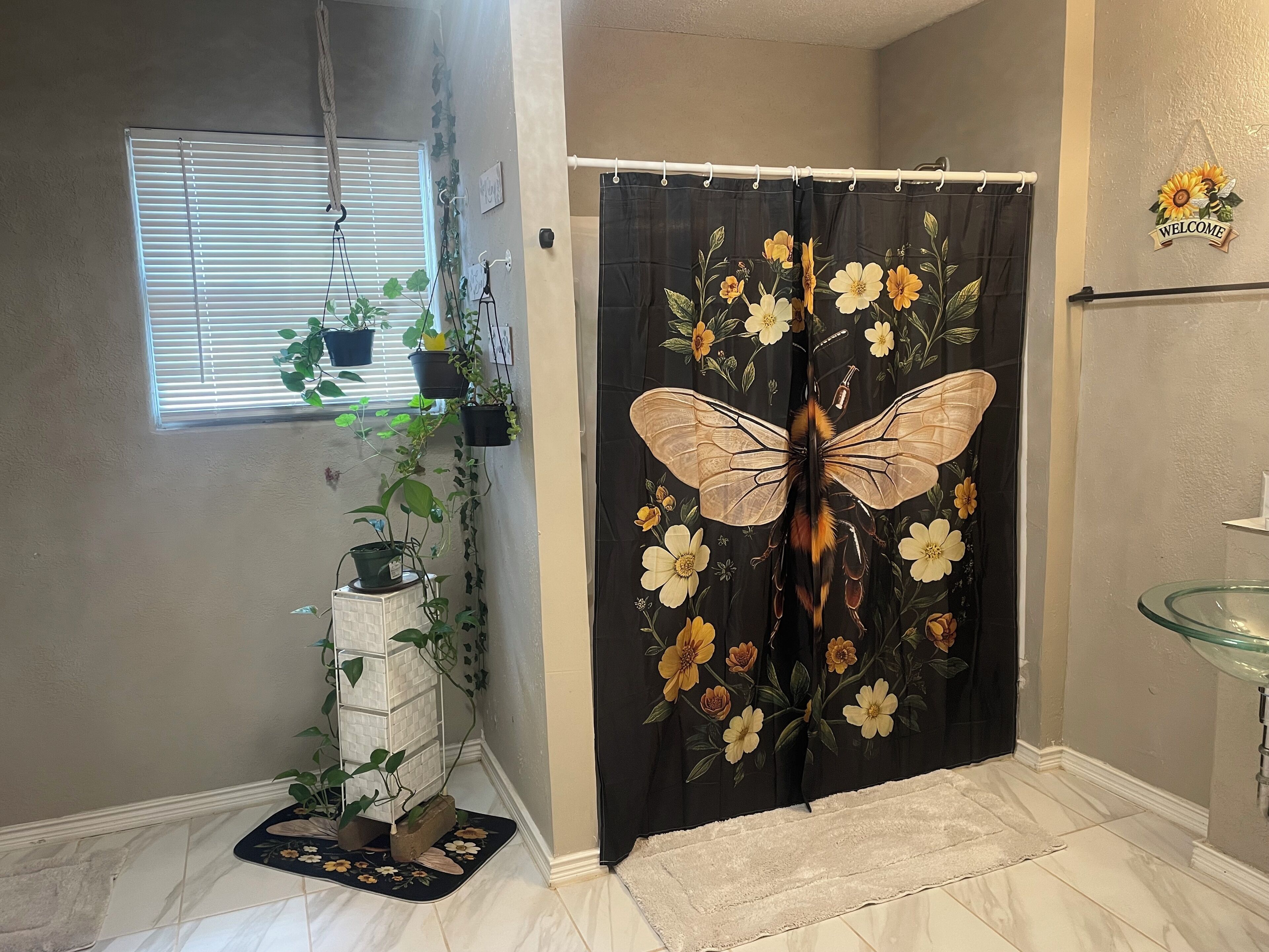 Un baño con cortina de ducha con diseño de mariposa, una ventana con persianas y una planta en maceta.