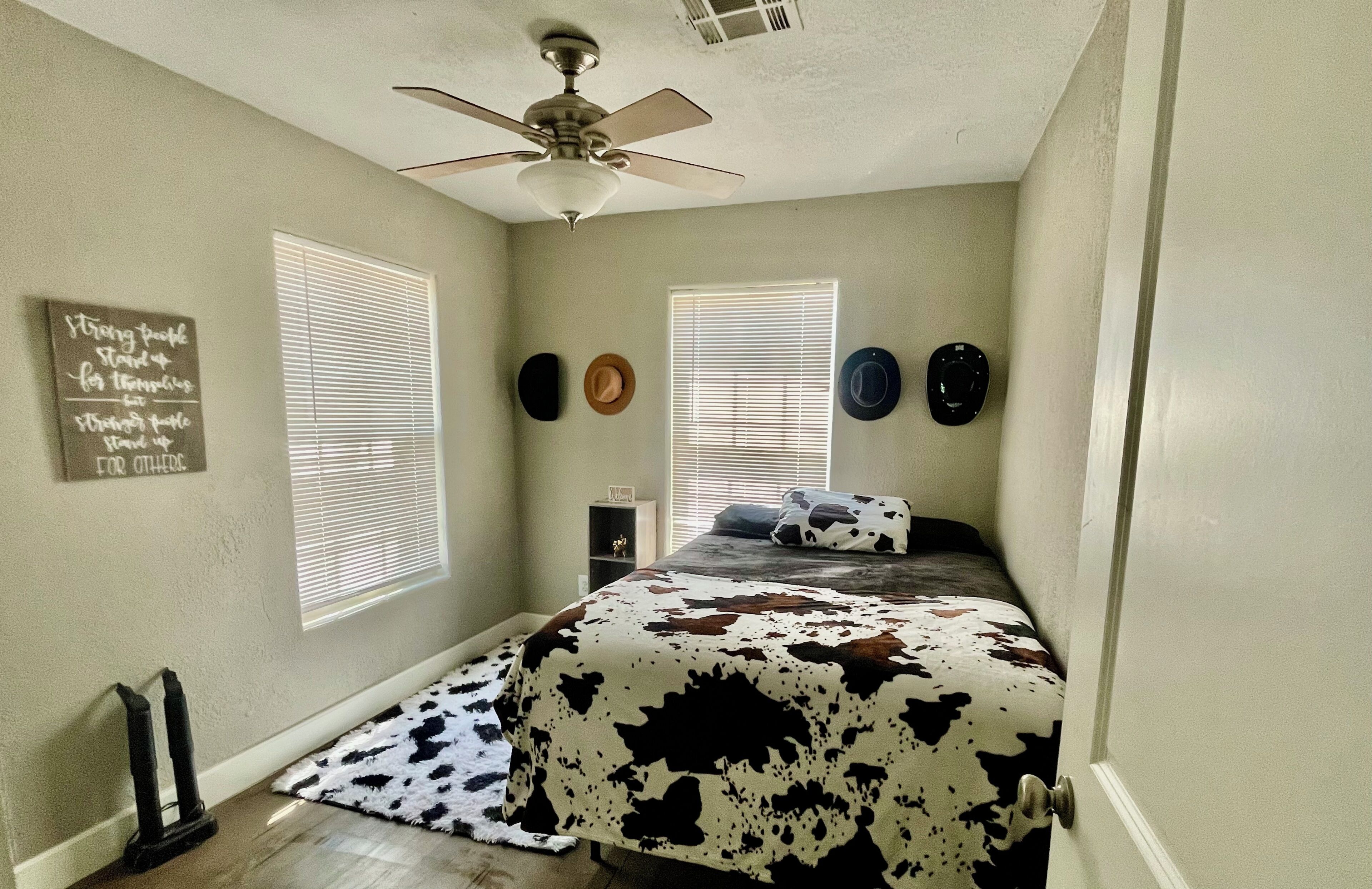 A bedroom with a bed, ceiling fan, two windows with blinds, a wall with decorative plates, and a small shelf.