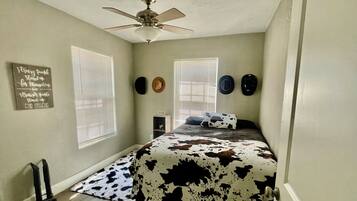 A bedroom with a bed, ceiling fan, two windows with blinds, a wall with decorative plates, and a small shelf.
