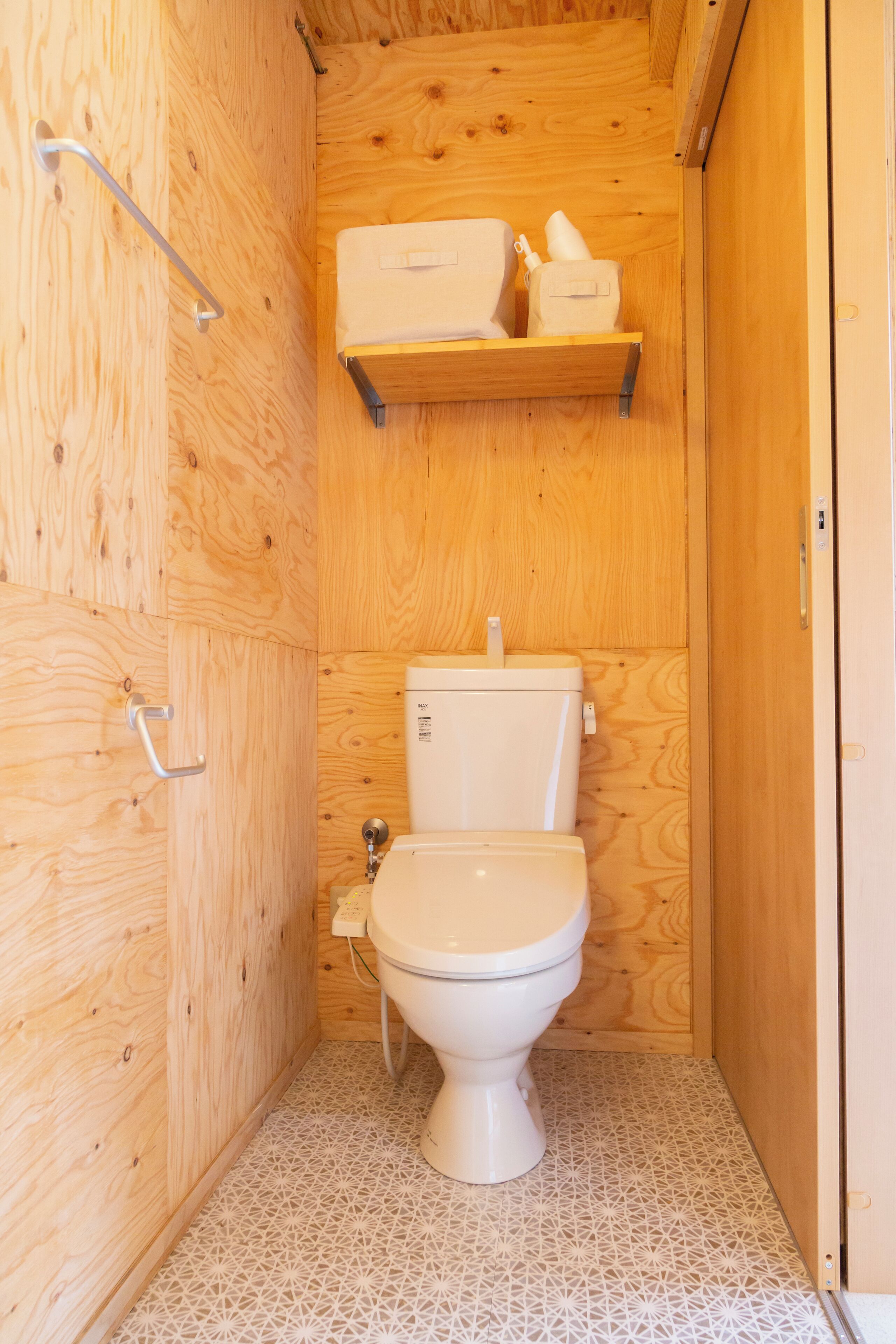 A small, wooden-paneled bathroom with a toilet, a shelf above the toilet, and a patterned floor.