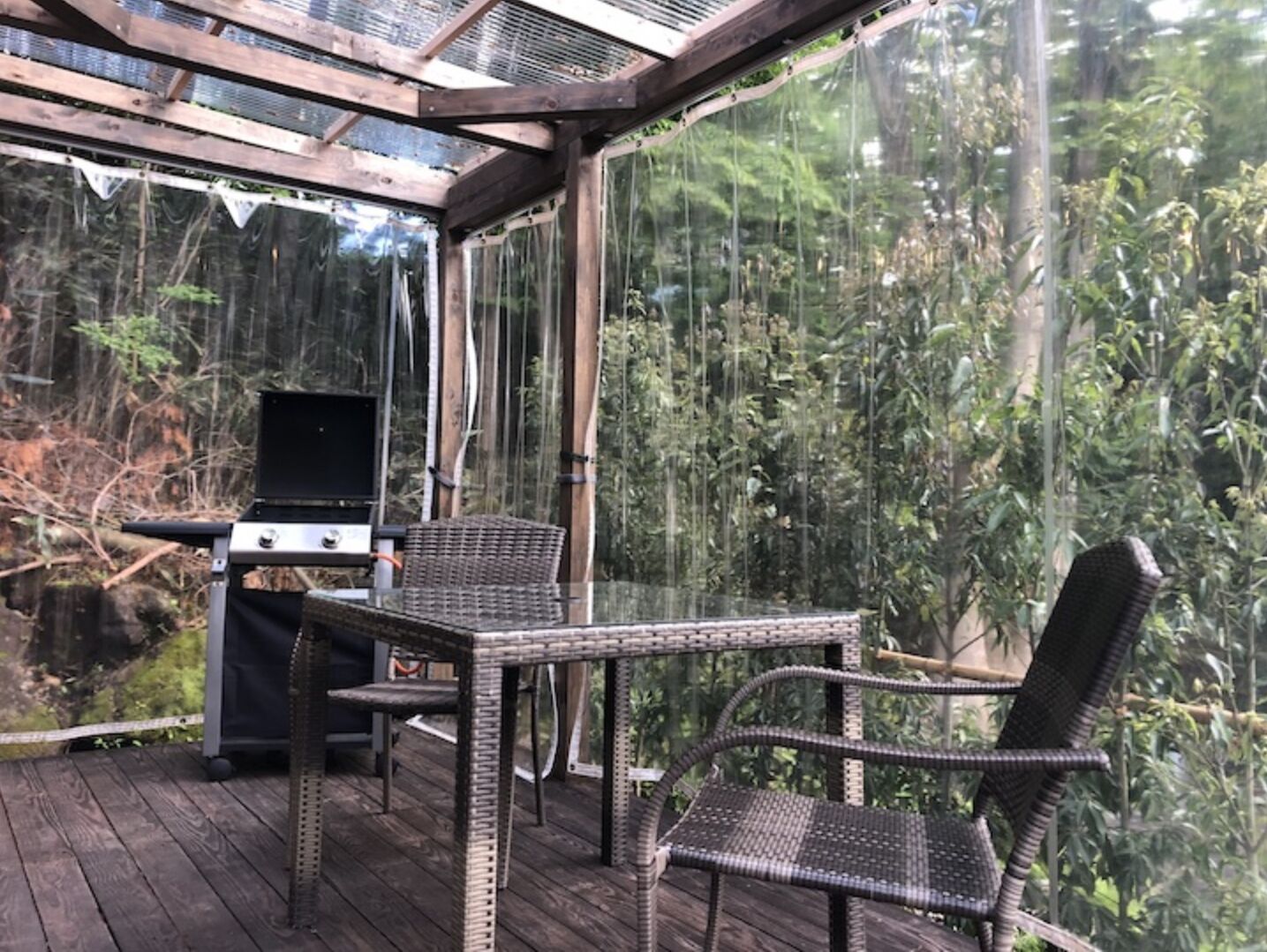 Une terrasse extérieure équipée d’un barbecue, de meubles en osier et d’une véranda en verre.
