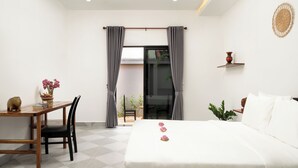 A hotel room with a bed, a desk, a chair, a small table with a vase of flowers, and a view through a glass door. - Visak Pool Villa  (Siem Reap)